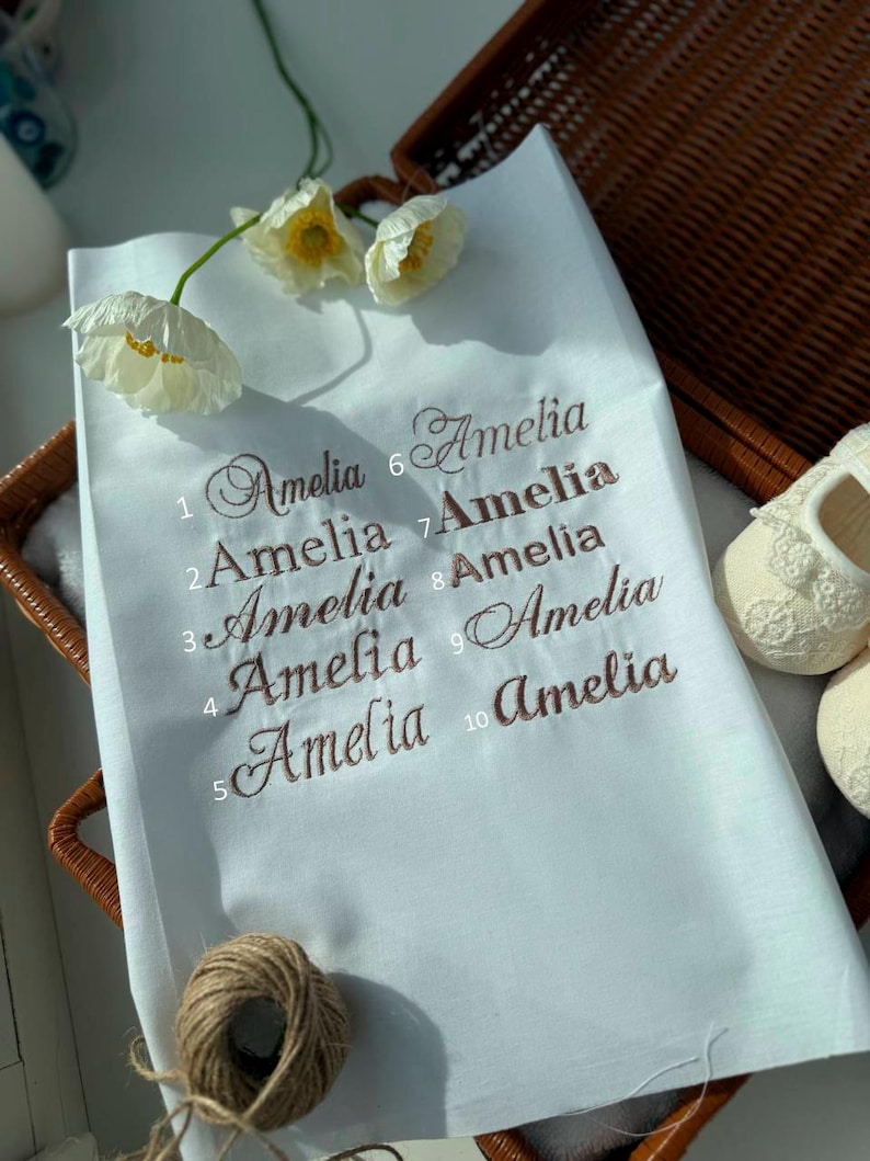 May include: White fabric with the name "Amelia" embroidered in brown script ten times. The fabric is in a wicker basket with white baby shoes and white flowers. A ball of twine is in the lower left corner.