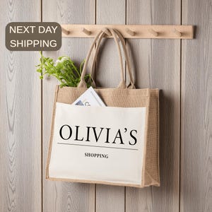 A beige tote bag with the text "OLIVIA'S SHOPPING" printed on it, hanging on a wooden hanger. The bag has a small plant in its pocket.