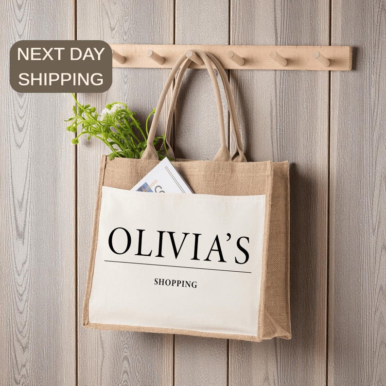 A beige tote bag with the text "OLIVIA'S SHOPPING" printed on it, hanging on a wooden hanger. The bag has a small plant in its pocket.
