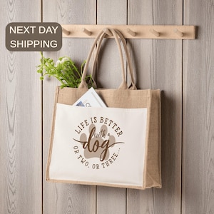 May include: A beige jute tote bag with a white front pocket. The pocket features the text "LIFE IS BETTER with a dog OR TWO, OR THREE..." in brown. The bag has long handles and is hanging on a wooden rack. The bag contains greenery and a magazine.