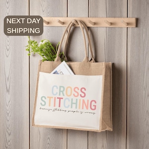 May include: A beige burlap tote bag with a white front pocket. The pocket features the words "CROSS STITCHING" in pastel colors, with the phrase "because stabbing people is wrong" below. The bag hangs on a wooden wall with a row of pegs.