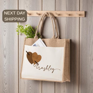 May include: A beige burlap tote bag with a white front pocket. The pocket features two brown yarn hearts pierced by a needle, with the name "Trashley" in cursive. The bag has beige handles and is hanging on a wooden rack.