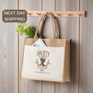May include: A beige burlap tote bag with a white front pocket featuring the text "SOCIETY of obstinate HEADSTRONG GIRLS". The bag has long handles and is hanging on a wooden rack. The bag contains greenery and a magazine.