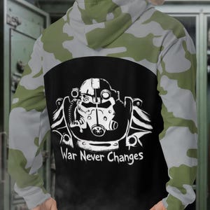 Könnte beinhalten: Ein Kapuzenpullover mit einem Tarnmuster in Grün- und Grautönen. Auf der Rückseite befindet sich ein schwarzes Feld mit einer weißen Grafik eines Helms und dem Text "War Never Changes".