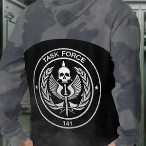 May include: A gray camouflage hoodie with a black panel on the back. The black panel features a white emblem with a skull, wings, and the text "TASK FORCE 141". The hoodie has a hood and long sleeves.