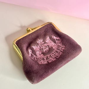 May include: A plum-coloured velvet coin purse with a gold-toned frame. The purse features the embroidered text "Juicy Couture" in pink, along with a crest design. The purse is open, revealing the interior.