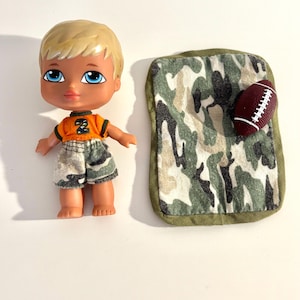 Könnte beinhalten: Eine Puppe mit blonden Haaren und blauen Augen, die ein orangefarbenes Oberteil mit der Nummer 2 und Camouflage-Shorts trägt. Eine Camouflage-Decke und ein weinroter Football liegen neben der Puppe.