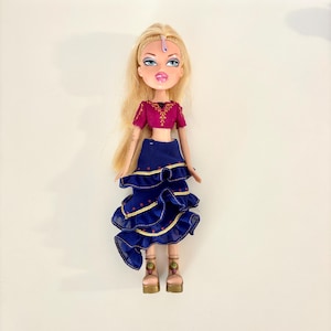 Può includere: Bambola bionda con occhi blu e labbra piene. Indossa un top corto magenta con ricami dorati e una gonna blu navy a balze con finiture dorate. La bambola è accessoriata con un copricapo e tacchi dorati.