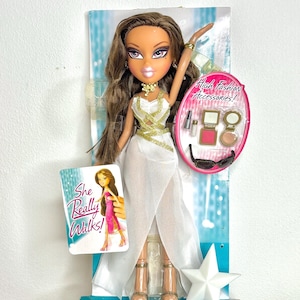 Può includere: Bambola Bratz in abito bianco con dettagli dorati, confezionata con accessori. La bambola ha lunghi capelli castani e tiene in mano una carta con la scritta "She Really Walks!". La confezione include il testo "High Fashion Accessories!"
