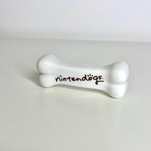 May include: A white, bone-shaped ceramic figurine with the text "nintendogs" in brown script. The figurine is a playful nod to the popular Nintendo game. The bone is smooth and glossy, with rounded ends and a slight curve.
