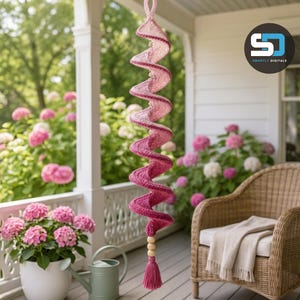 May include: A hanging spiral decoration crafted from yarn in an ombre gradient of pink and magenta. The structure features a twisted, corkscrew design with a loop at the top for suspension and a decorative wooden bead accent above a yarn tassel.