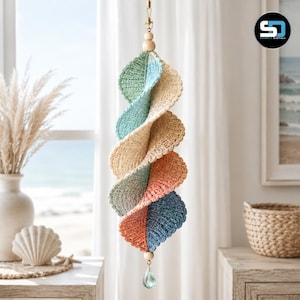Op de afbeelding: Een hangende spiraaldecoratie met gehaakte bladvormige segmenten in afwisselende tinten saliegroen, beige, koraal en blauw. Het object hangt aan een houten kraal en is aan de onderzijde afgewerkt met een heldere, gefacetteerde kristallen druppel.