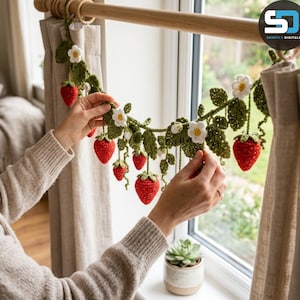 Pode incluir: Uma guirlanda de crochê com morangos vermelhos, folhas verdes e flores brancas com centros amarelos. O cordão decorativo está suspenso horizontalmente, exibindo vários elementos de morango e flores feitos de fio, pendurados em uma haste de madeira.