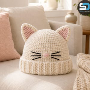 May include: A cream-coloured crochet beanie featuring two pointed cat ears with pink inner detailing. The front of the hat displays a small pink triangular nose and six black embroidered whiskers arranged in two sets of three on either side.