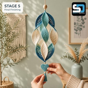 K&ouml;nnte beinhalten: Ein h&auml;ngendes Mobile mit einer Spirale aus geh&auml;kelten Blattformen in Blau-, T&uuml;rkis- und Cremet&ouml;nen. Die Struktur ist mit kleinen Holzperlen und einem zentralen geh&auml;kelten Herz an der Unterseite verziert, das mit einer blauen Quaste endet.