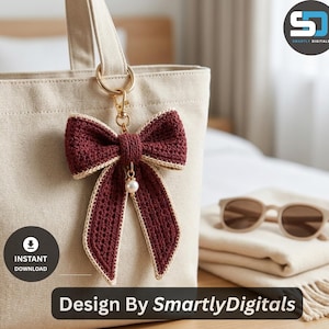 May include: A dark maroon crochet bow keychain with a cream-colored border and a small white pearl charm attached to a gold-toned metal lobster clasp and ring, hanging from a beige canvas tote bag with the text Design By SmartlyDigitals.