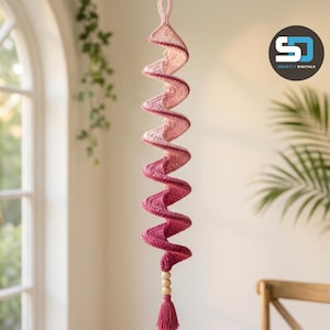 May include: A hanging spiral decoration made of crocheted yarn in an ombre gradient, transitioning from light pink at the top to deep magenta at the bottom. The piece features a loop for hanging, three wooden beads, and a matching yarn tassel.