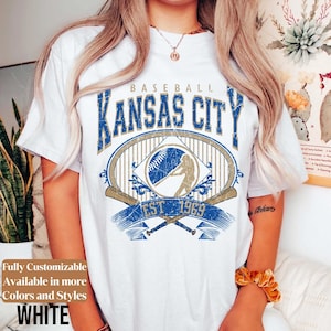 May include: White t-shirt featuring a distressed blue and gold graphic design. The design includes the text "KANSAS CITY BASEBALL EST 1969" with a baseball and batter illustration. The shirt is fully customizable and available in more colors and styles.