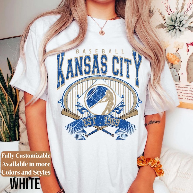 May include: White t-shirt featuring a distressed blue and gold graphic design. The design includes the text "KANSAS CITY BASEBALL EST 1969" with a baseball and batter illustration. The shirt is fully customizable and available in more colors and styles.