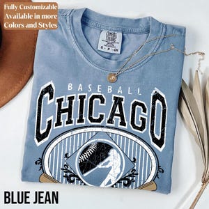 May include: A blue jean colored t-shirt with the word "CHICAGO" in large black letters. The shirt features a baseball design with a batter silhouette and baseball graphic. The text "BASEBALL" is above the word "CHICAGO". The shirt is a Comfort Colors brand.