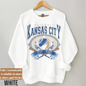 May include: White sweatshirt with a baseball-themed graphic. The design features the text "BASEBALL KANSAS CITY EST 1969" in blue and gold, along with baseball bats and a baseball. The sweatshirt is hanging on a wooden hanger.