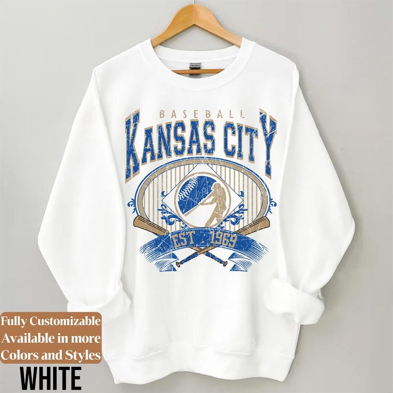 May include: White sweatshirt with a baseball-themed graphic. The design features the text "BASEBALL KANSAS CITY EST 1969" in blue and gold, along with baseball bats and a baseball. The sweatshirt is hanging on a wooden hanger.