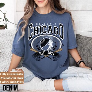 May include: Denim blue t-shirt with a baseball graphic. The design features the word "Chicago" in a bold font, a baseball, a batter silhouette, crossed bats, and the text "EST. 1894". The shirt has short sleeves.