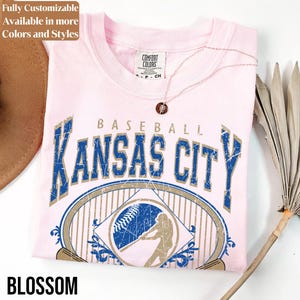 May include: A light pink t-shirt with a distressed "Kansas City Baseball" graphic in blue and tan. The shirt has a crew neck and short sleeves. The word "Blossom" is printed at the bottom. A necklace with a pendant is on the shirt.