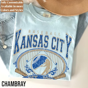 May include: Light blue t-shirt with a distressed graphic that says "Baseball Kansas City" in blue and tan. The shirt features a baseball and a batter illustration. The word "Chambray" is at the bottom.