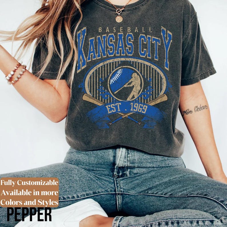 May include: Dark gray baseball-themed t-shirt with "Kansas City" in blue and gold lettering. The shirt features a baseball player graphic, crossed bats, and "EST. 1969". The text "Fully Customizable Available in more Colors and Styles PEPPER" is also visible.