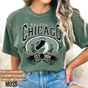 May include: Green t-shirt with a baseball-themed graphic. The design features the word "Chicago" in a bold font, a baseball, a batter silhouette, and the text "EST 1894". The shirt is a moss green color.