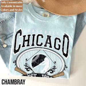 May include: A light blue t-shirt featuring the text CHICAGO in large black distressed lettering above a graphic of a baseball diamond, a baseball, and a batter. The shirt has a Comfort Colors label at the inner neckline.