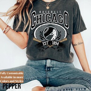 May include: Dark gray baseball t-shirt with a distressed graphic. The graphic features the word "CHICAGO" in a bold font, a baseball player silhouette, and crossed bats. The text "EST. 1894" is also included. The shirt also has the text "Fully Customizable Available in more Colors and Styles PEPPER".