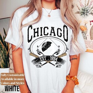 May include: White t-shirt with a black and white graphic design featuring the word "Chicago" above a baseball and crossed bats. The text "EST 1894" is below the baseball graphic. The shirt also has the text "Fully Customizable Available in more Colors and Styles WHITE".