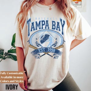 May include: Ivory-colored t-shirt featuring a distressed baseball design. The design includes the words "TAMPA BAY" in blue, a baseball, crossed bats, and the text "EST 1998". The shirt is fully customizable and available in more colors and styles.