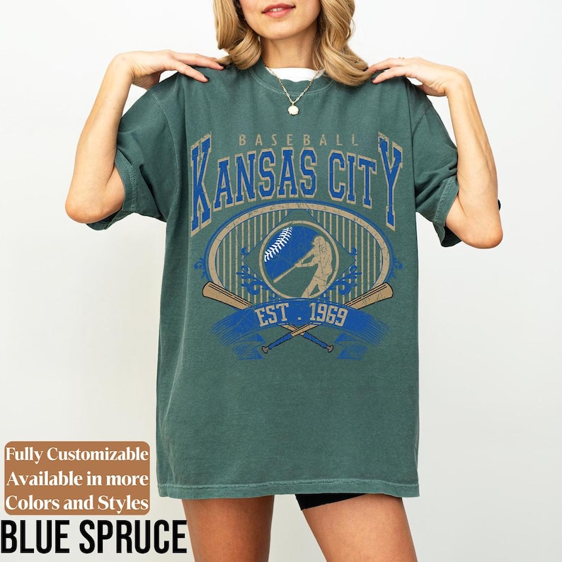 May include: Blue spruce baseball t-shirt with "Kansas City" in blue and gold lettering. The shirt features a baseball graphic with a batter and crossed bats, and the text "EST 1969". The shirt is fully customizable and available in more colors and styles.