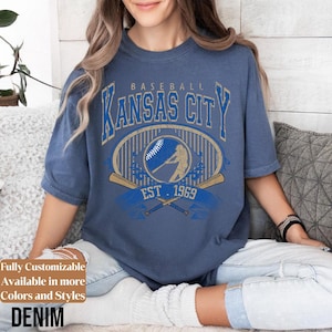 May include: Denim blue t-shirt with a vintage baseball graphic. The design features "Kansas City" in a bold, arched font, a baseball, and crossed bats, with "EST. 1969". The shirt is a relaxed fit.