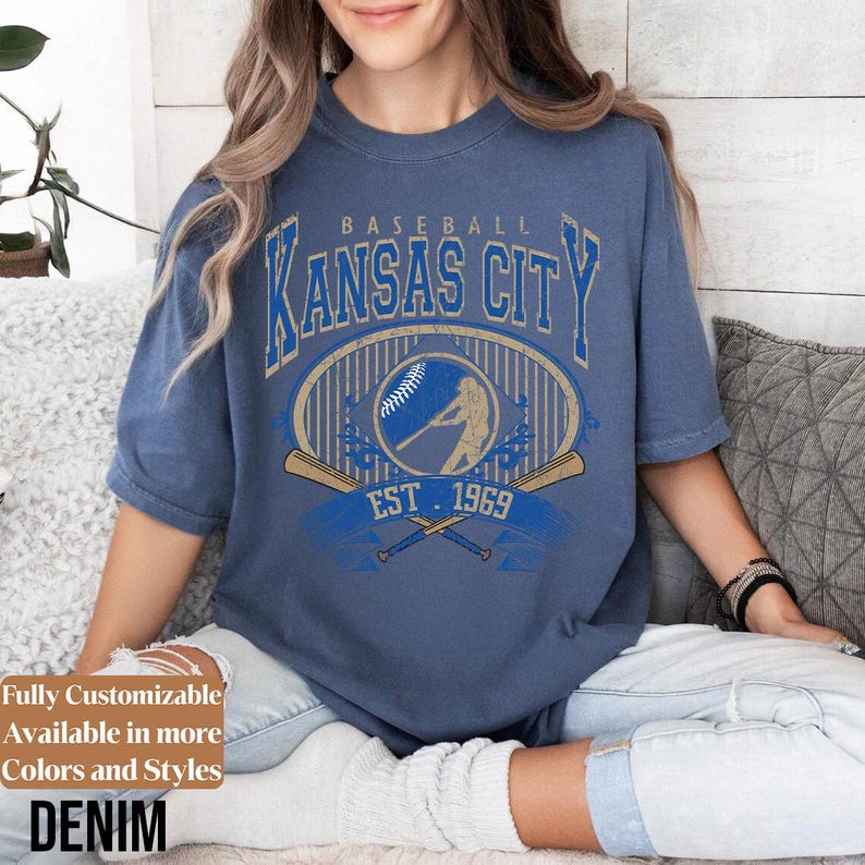 May include: Denim blue t-shirt with a vintage baseball graphic. The design features "Kansas City" in a bold, arched font, a baseball, and crossed bats, with "EST. 1969". The shirt is a relaxed fit.