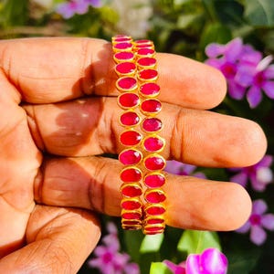 Puede incluir: Dos brazaletes circulares que presentan una serie de piedras rojas ovaladas engastadas en un marco de metal dorado. Cada piedra está engastada individualmente, creando una fila continua de gemas rojas alrededor de la circunferencia de ambos.