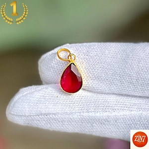 Pode incluir: Um pingente em forma de gota com uma pedra preciosa vermelha escura facetada, engastada numa moldura de metal dourado. O pingente inclui uma pequena argola circular dourada no topo para fixação, apresentando um acabamento polido e vivo.