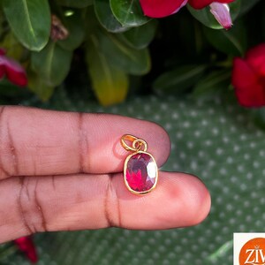 May include: A small oval-shaped pendant featuring a faceted deep red gemstone set in a polished gold-toned metal frame. The pendant includes a simple gold-toned bail at the top for attachment to a chain, showcasing a clear, reflective surface.