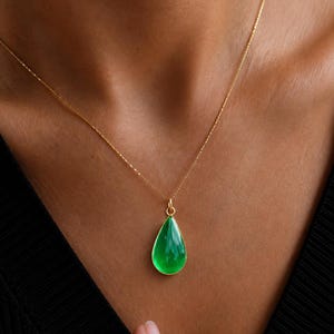Puede incluir: Un collar de cadena dorada con un colgante en forma de lágrima que presenta una piedra verde lisa y vibrante engastada en un bisel dorado. El colgante cuelga de una delicada cadena de eslabones dorados, creando una pieza de joyería elegante.