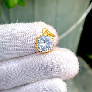 Puede incluir: Un colgante redondo con una piedra preciosa transparente y facetada engastada en un bisel de tono dorado pulido. El colgante incluye una pequeña anilla circular de tono dorado en la parte superior, con una superficie brillante y reflectante.