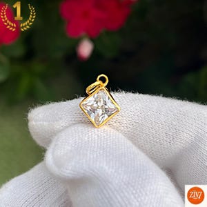Puede incluir: Un pequeño colgante de piedra preciosa transparente de corte cuadrado engastado en un marco de metal dorado. El colgante presenta una superficie facetada que refleja la luz e incluye una pequeña anilla circular dorada en la parte superior.