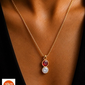May include: A gold-tone chain necklace featuring a vertical pendant with two circular bezel-set stones. The top stone is a vibrant red, while the bottom stone is a clear, faceted white gem. The pendant hangs from a delicate, thin gold-coloured link chain.