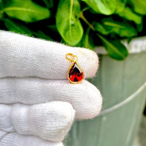 Puede incluir: Un colgante en forma de lágrima con una piedra preciosa facetada de color rojo intenso engastada en un marco de metal dorado pulido. El colgante incluye una pequeña anilla circular dorada en la parte superior para sujetarlo a una cadena.
