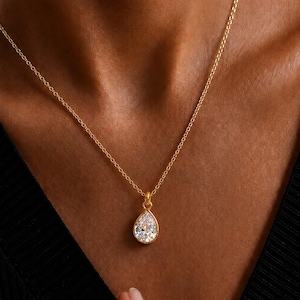 Puede incluir: Un collar de cadena dorada con un colgante de piedra transparente en forma de pera engastado en un bisel dorado. El colgante cuelga de una delicada cadena de eslabones dorados. El diseño es sencillo y destaca la piedra facetada y el acabado.