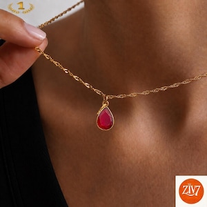 Könnte beinhalten: Eine goldfarbene gedrehte Kette mit einem einzelnen tropfenförmigen Anhänger mit einem tiefroten Edelstein. Der Anhänger hängt an einer zarten Gliederkette, die auf der Haut aufliegt. Ein kleines goldenes Symbol mit der Nummer eins ist zu sehen.