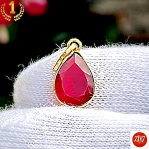 May include: A teardrop-shaped pendant featuring a faceted deep red gemstone set in a gold-toned metal frame. The pendant includes a small circular bail at the top for attachment, showcasing a polished finish and a vibrant, saturated red colour.