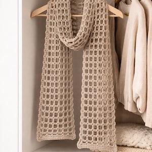 May include: A beige, open-weave crochet scarf hangs on a wooden hanger in a wardrobe. The scarf has a rectangular pattern and is draped over the hanger. Other beige clothing items are visible in the background.